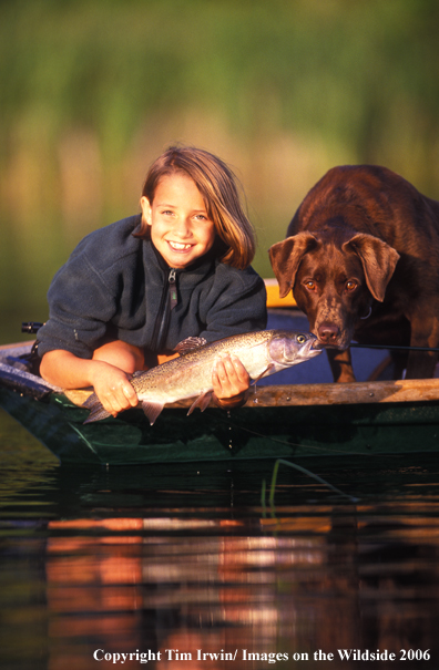 Chocolate Labrador Retriever in boat with young girl holidng a trout.