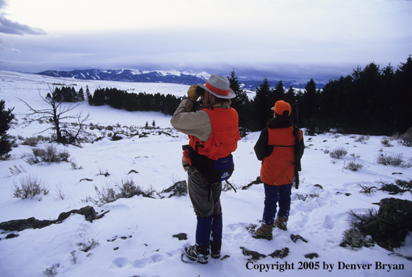 Father and son hunters glassing for big game in a field in winter.