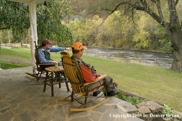 Flyfishermen talking on the porch of a fishing club house overlooking a creek.